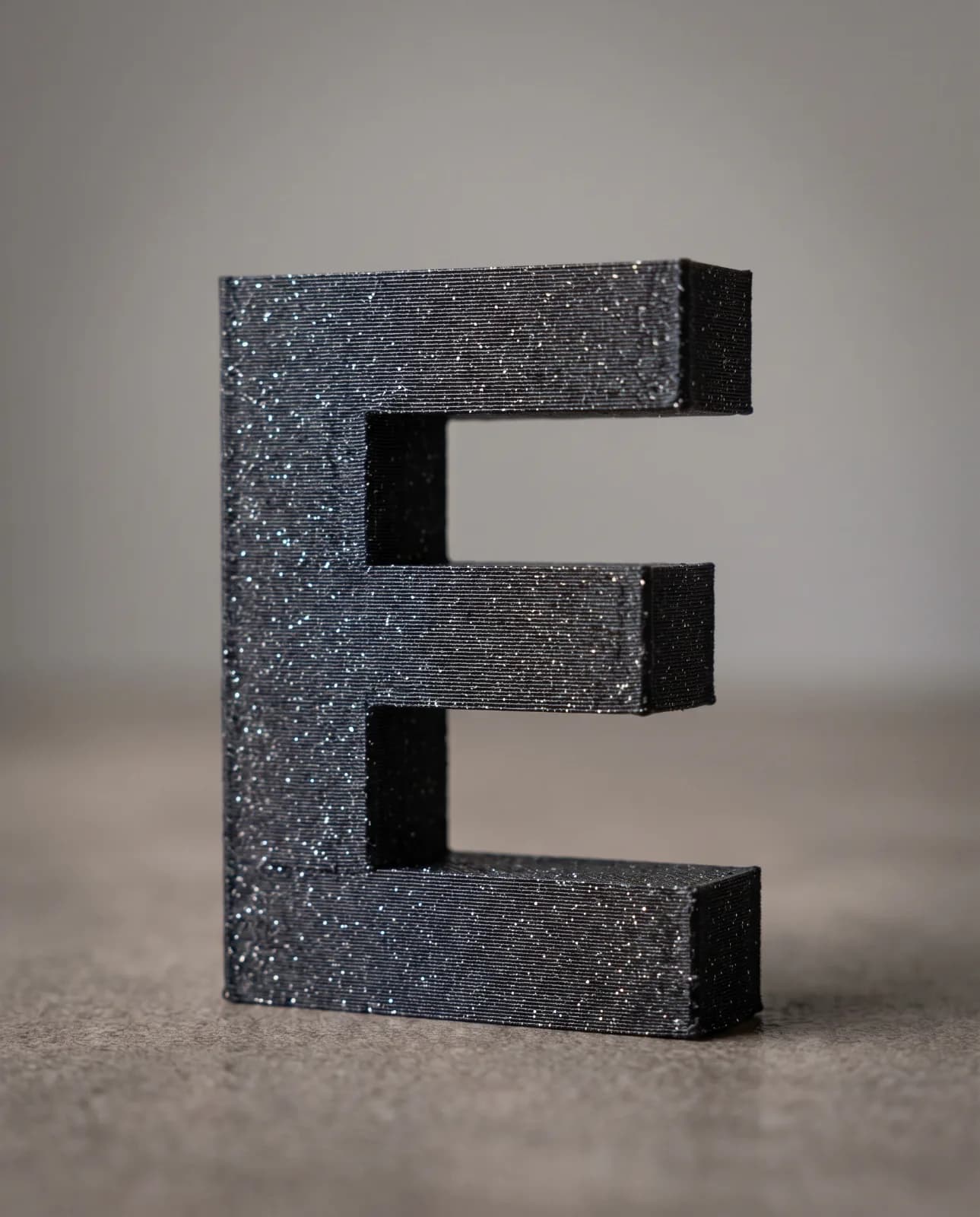 A black 3D-printed letter E in glitter filament, flecked with sparkle