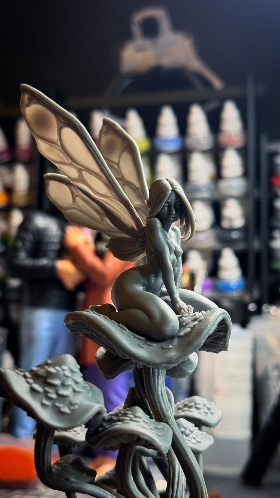 An unpainted resin print of a fairy sitting on a cluster of mushrooms, in front of the studio paint rack