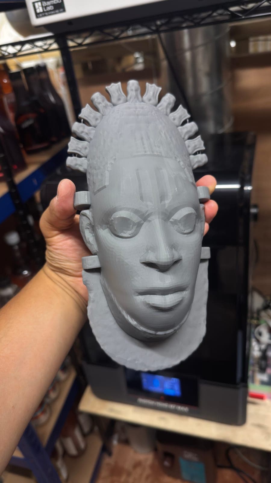 A large unpainted resin print of a carved mask, held in front of the resin printer