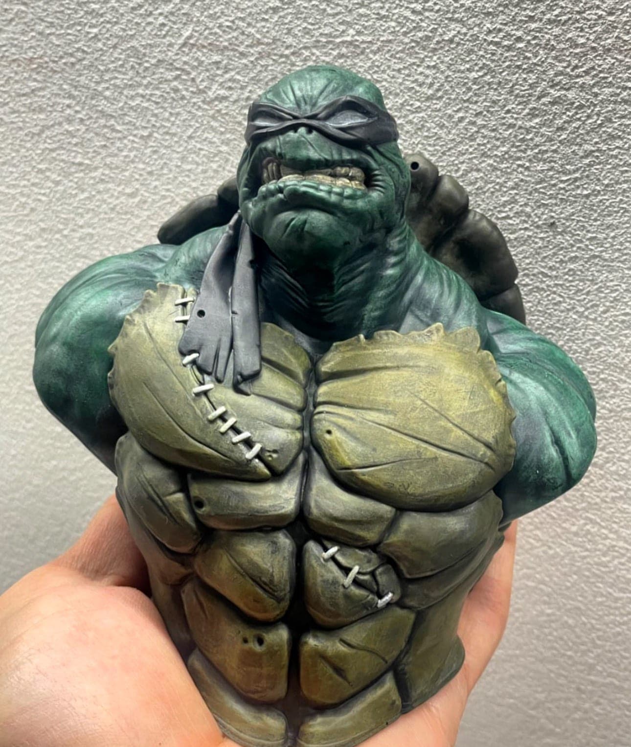 A hand-painted resin bust of a green masked brute with a stitched chest, held in the hand