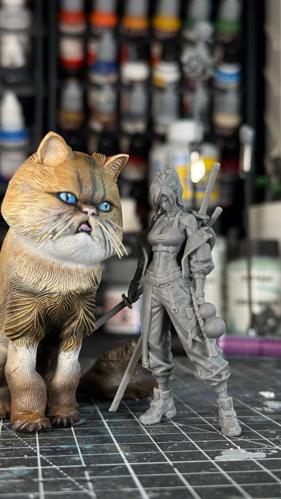 A hand-painted cat figure beside an unpainted resin figure, in front of the paint rack