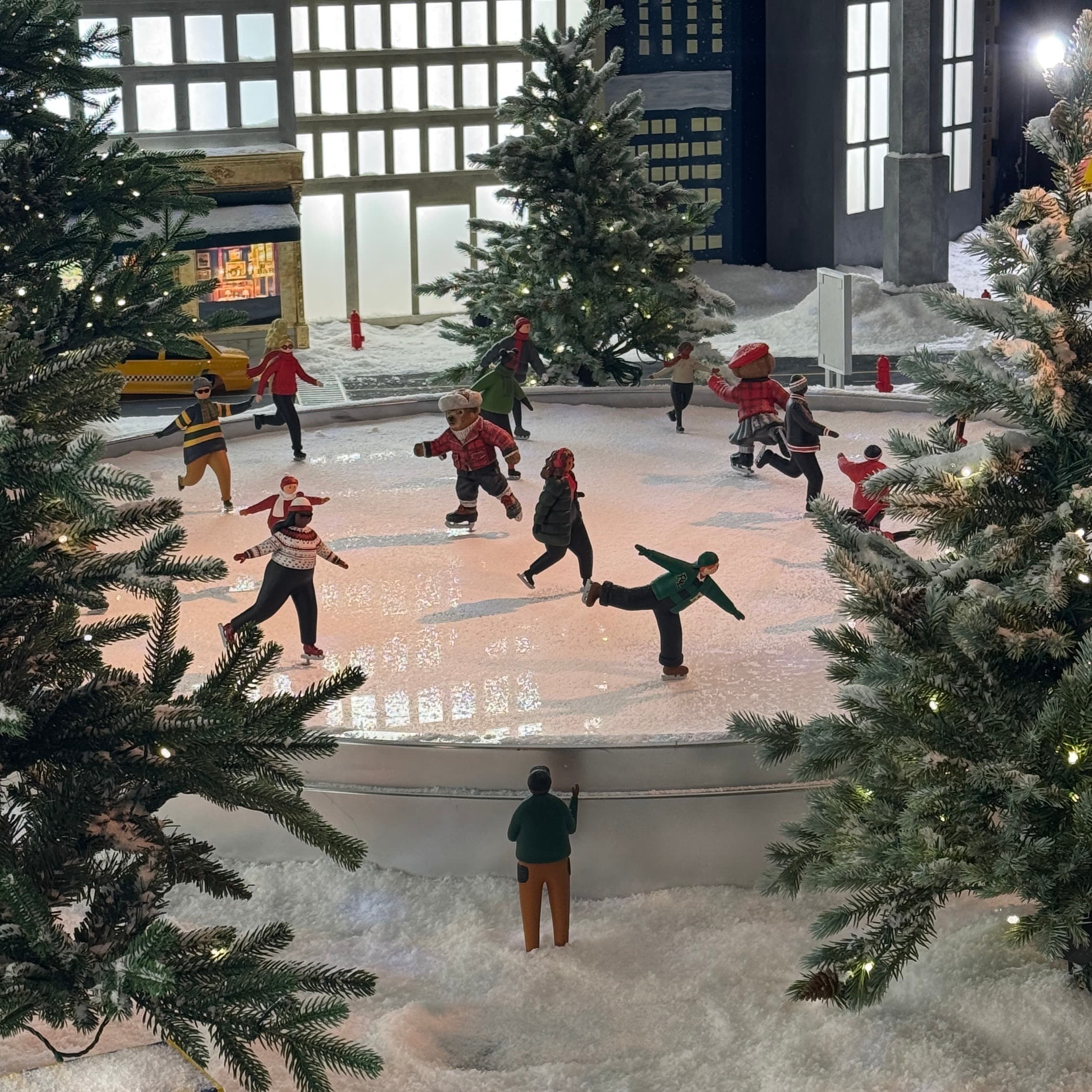 Miniature skaters and bears fill an ice rink surrounded by snow-covered fir trees, lights and model buildings.